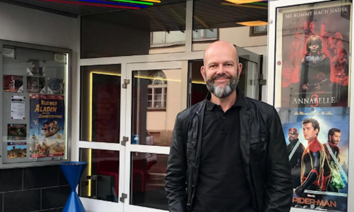 Ein lächelnder Mann mit dunkler Kleidung vor einem Kinoeingang. Am Engang hängen verschiedenen Filmplakate