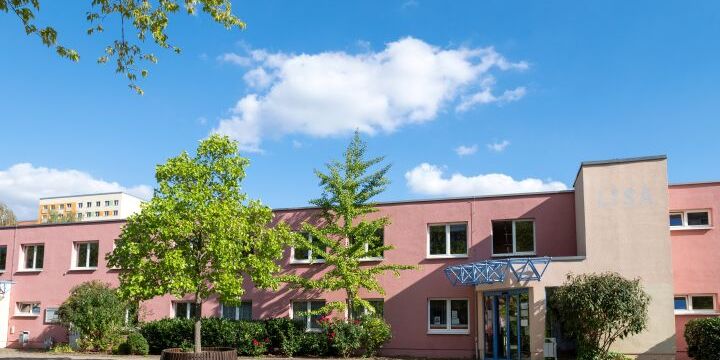 Ein rosarabenes langezogenes Gebäude mit zwei Stockwerken. Davor steht ein Baum mit einer Sitzbank, die den Stamm umrundet.  &copy;C. Worsch